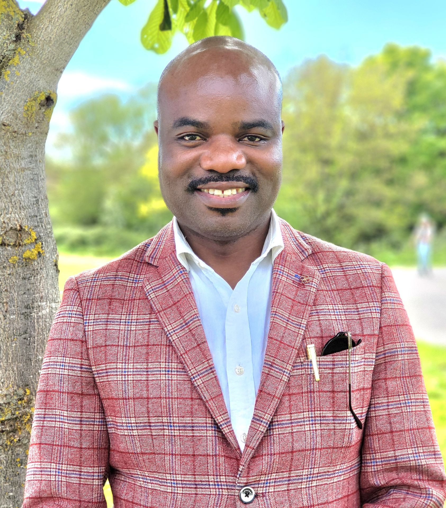 Smiling person in a red plaid jacket stands by a tree in a green outdoor setting.